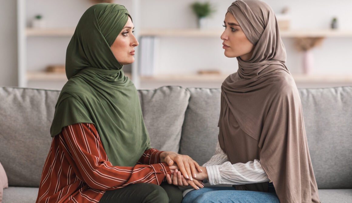 Unhappy Mature Muslim Mother And Adult Daughter Holding Hands Indoor Unhappy Mature Muslim Mother And Adult Daughter Sitting Holding Hands Supporting Each Other At Home, Wearing Hijab. Middle-Eastern Family Problems And Struggles Concept